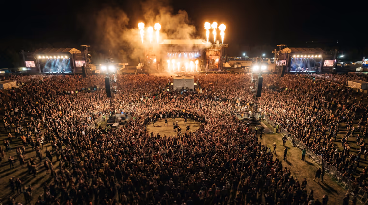 Vue aérienne d'un festival metal de nuit, scènes monumentales et pyrotechnie au-dessus de la foule